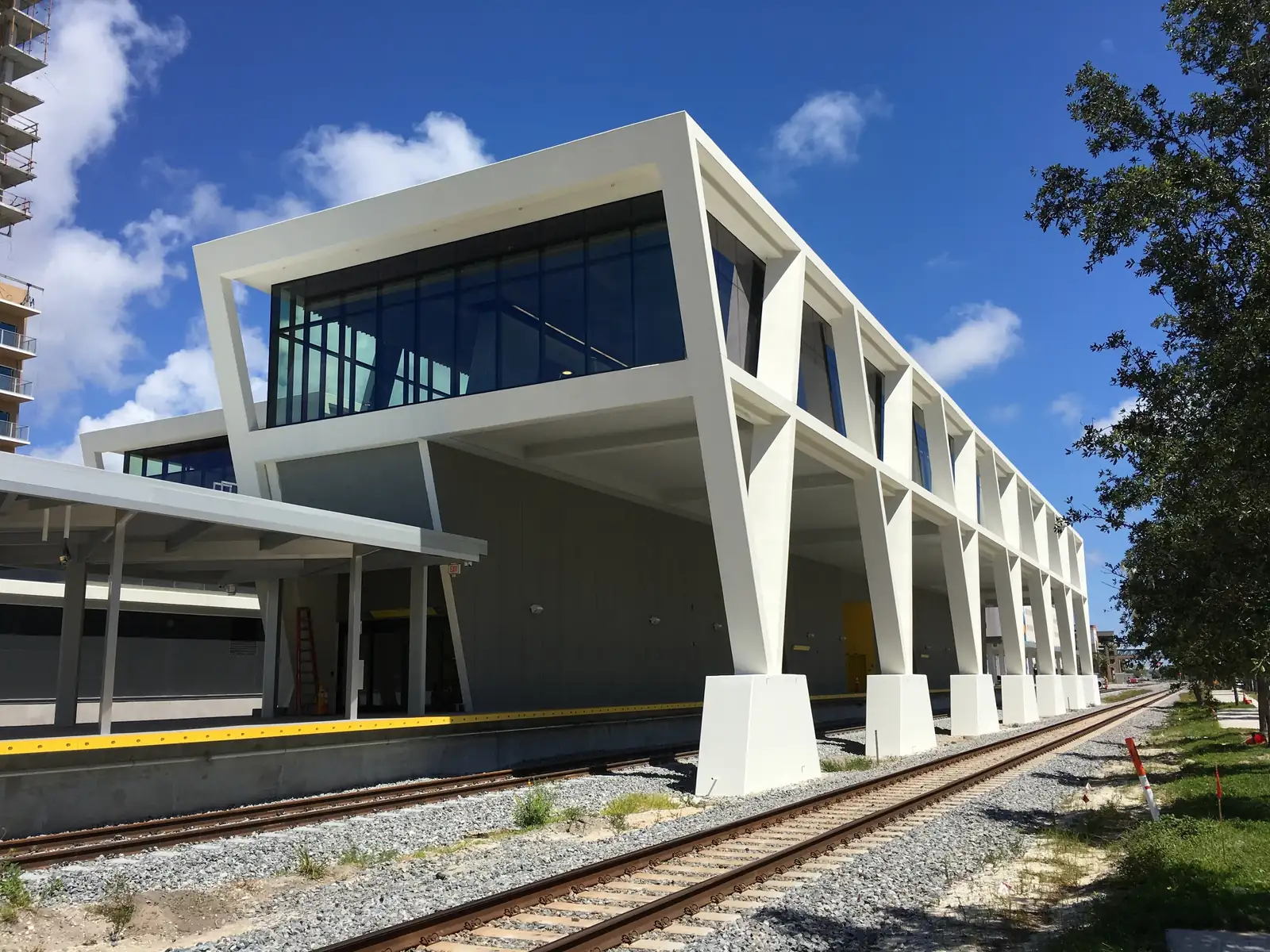 The white Brightline station seen from beside the tracks: a long elevated glass box carried on angled white V-shaped piers over the platform, ballast and rails in the foreground, a tree at right and a blue sky with white clouds.