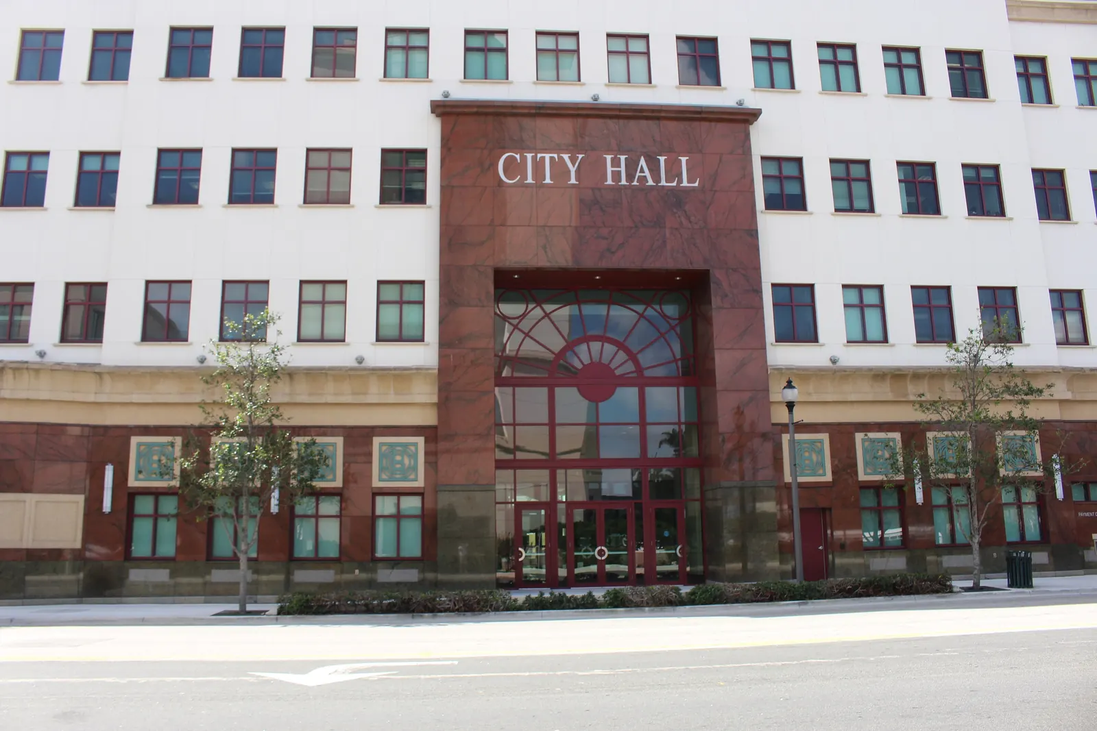 Straight-on view of the City Hall entrance: a tall red-granite portal with 'CITY HALL' lettering and an arched fan window over glass doors, set in a cream multi-storey facade with rows of windows, young trees and a street lamp on the sidewalk, and an empty road in front.