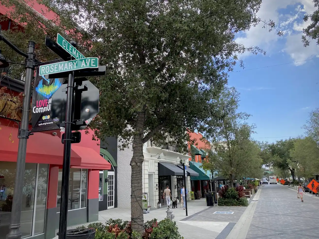 Street-sign post reading Clematis St and Rosemary Ave in the foreground, a shade tree, red awnings and shopfronts at left, and a wide brick-paved sidewalk receding past cafes and pedestrians under a blue sky with white clouds.
