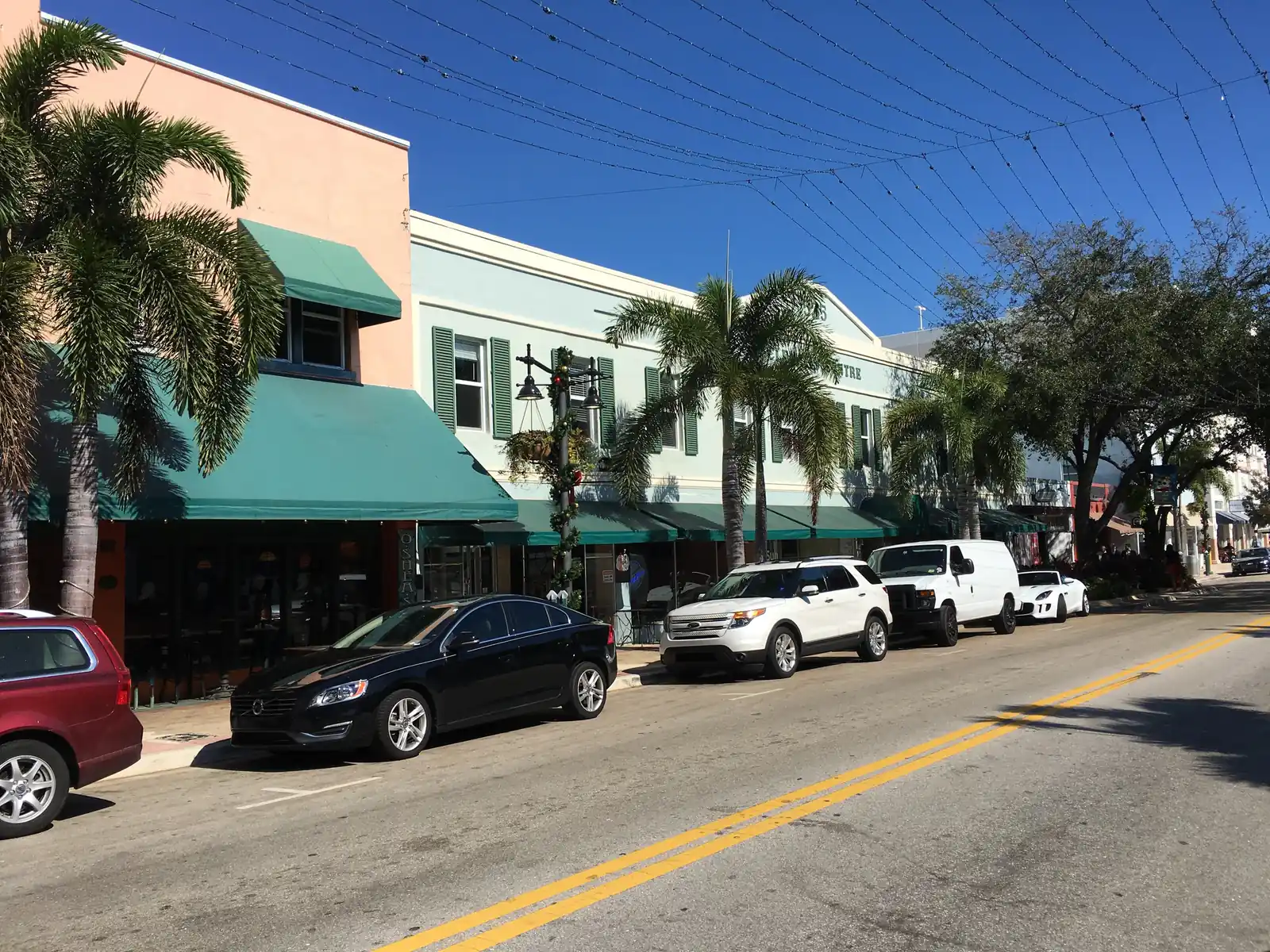 Sunny day on Clematis Street: a row of restored two-storey shopfronts in salmon, mint and cream with long green awnings, palms and shade trees along the sidewalk, cars parked at the kerb, overhead string-light wires and a deep blue sky.
