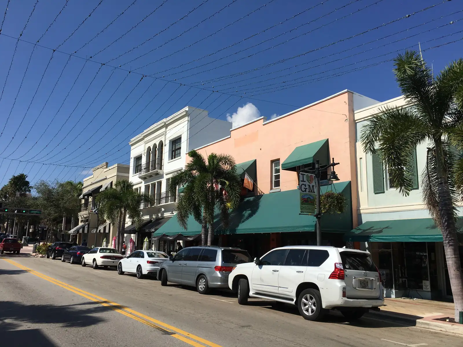 Clematis Street under a cloudless blue sky criss-crossed by overhead string-light wires: pastel two-storey historic buildings with green awnings and palms on the right, a line of parked cars and SUVs along the kerb, yellow centre lines in the foreground.