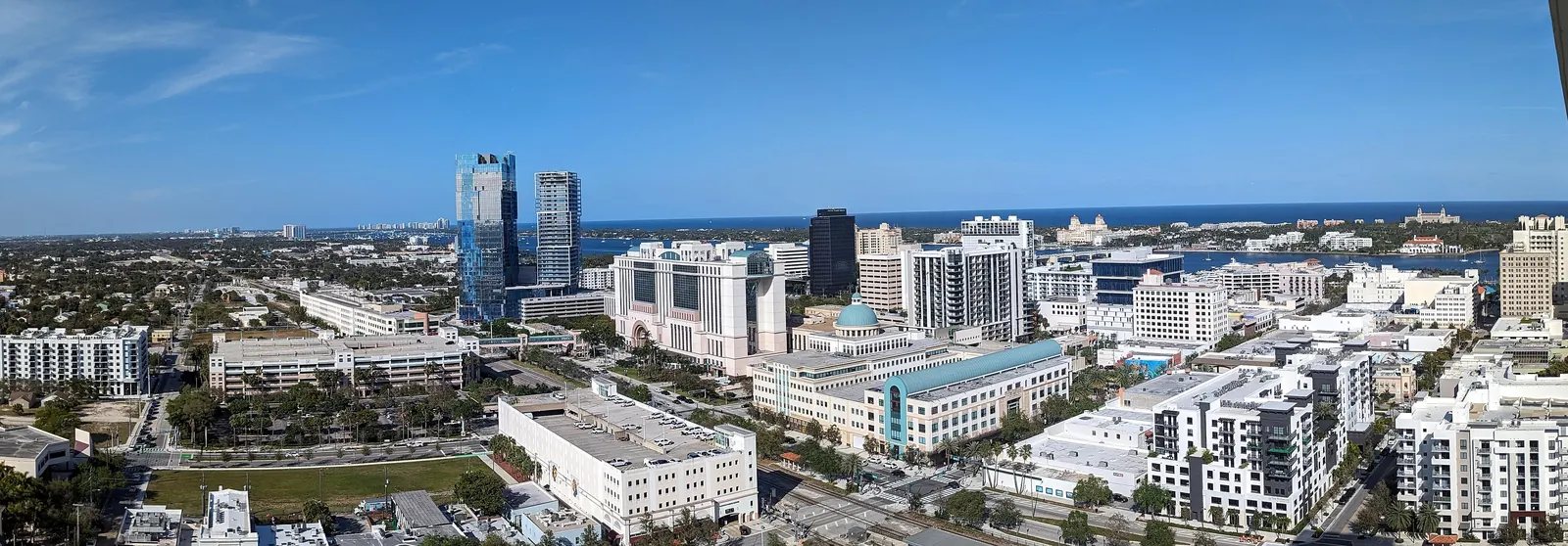 High rooftop panorama over downtown West Palm Beach on a clear day: two new glass towers at left, a large pink civic complex and a green-domed building at centre, new white apartment blocks at right, rail tracks below, and the blue lagoon, Palm Beach island and the ocean on the horizon.