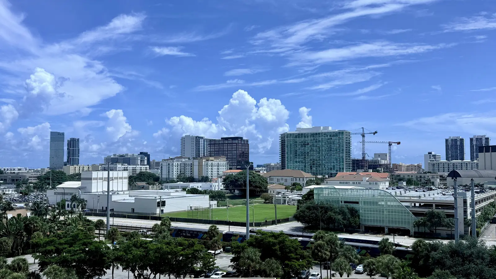 Summer view over treetops and a low green-glass building beside a grassy field to the downtown skyline: glass office towers, a tan high-rise at right and construction cranes, under a blue sky with tall white cumulus clouds.