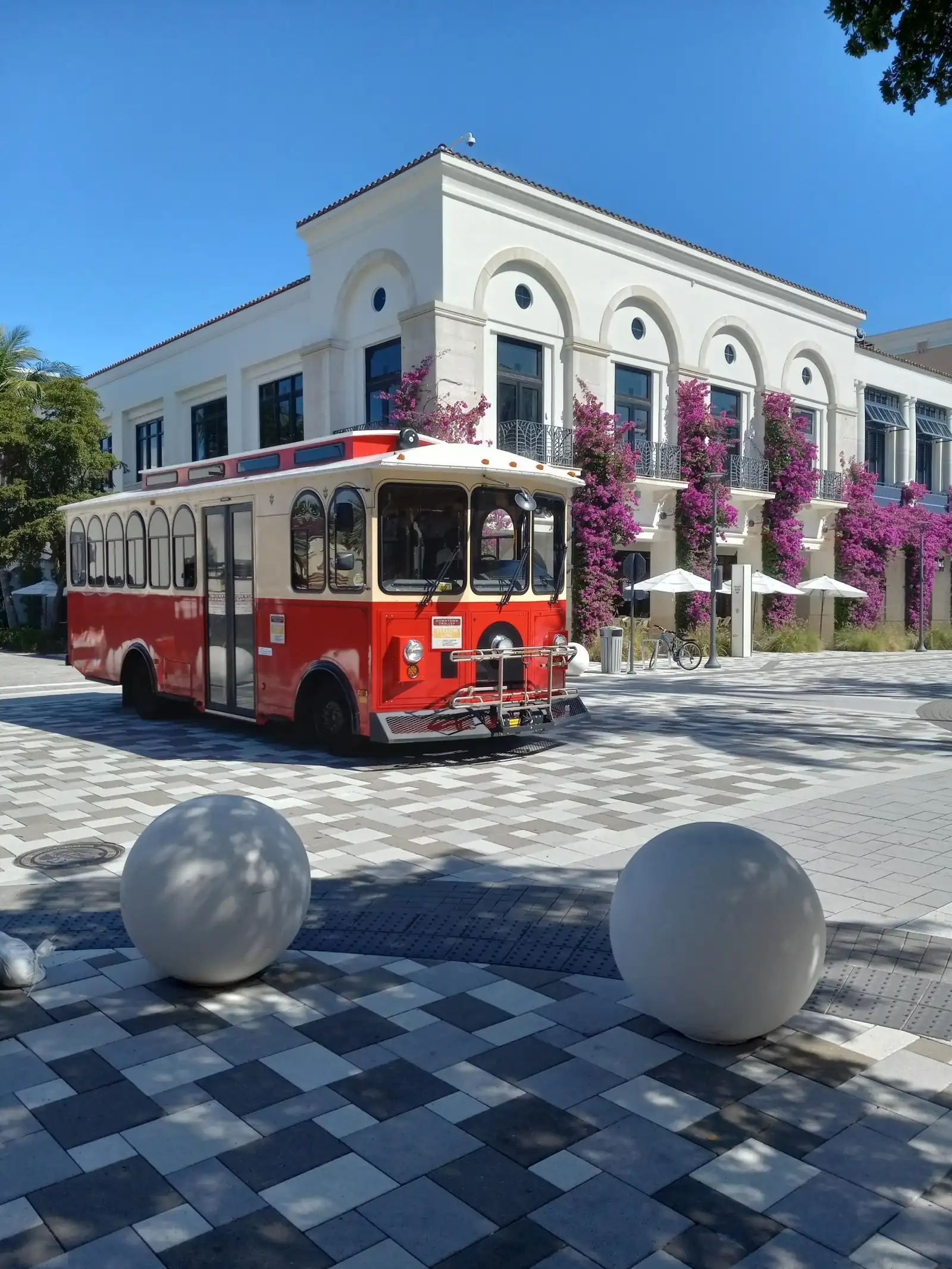 Red and cream downtown trolley bus parked on a patterned grey paver plaza beside two large white stone spheres, in front of a white arched building draped in magenta bougainvillea, under a clear blue sky.