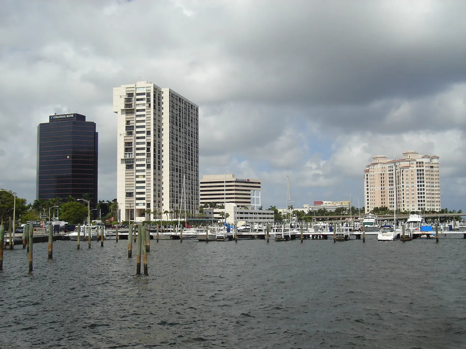 Overcast view across grey water and rows of marina pilings to moored boats, a dark glass office tower, a tall white residential tower and, further south, a cream high-rise on the waterfront.