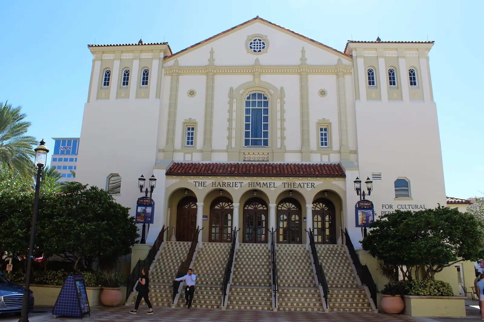 Front of the cream Mediterranean-revival former church, now the Harriet Himmel Theater: twin tower wings, a rose window, five arched doorways under a red-tiled porch roof bearing the theater name, and wide tiled stairs flanked by shrubs, lanterns and potted plants.