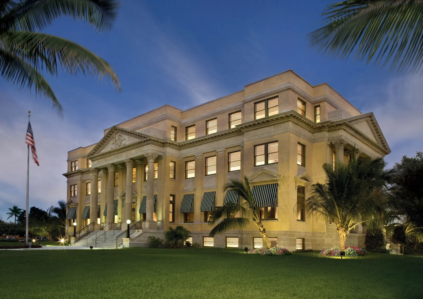 The restored 1916 neoclassical courthouse at dusk: a cream stone facade with a columned portico and pediment, green-striped awnings, warm lights in every window, a flag on a tall pole at left and a smooth green lawn in front under a deep blue sky.