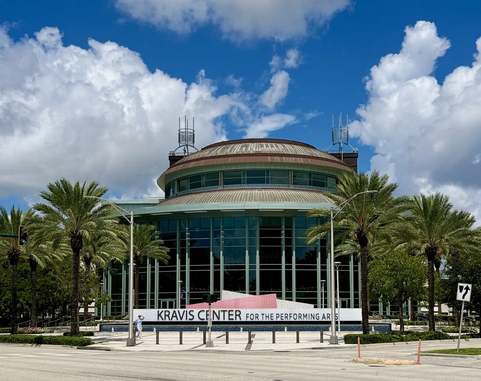 The Kravis Center's round glass-walled lobby drum under a domed roof, with a 'Kravis Center for the Performing Arts' sign on a low wall, royal palms and lamp posts, and a wide paved forecourt below a bright sky full of white clouds.
