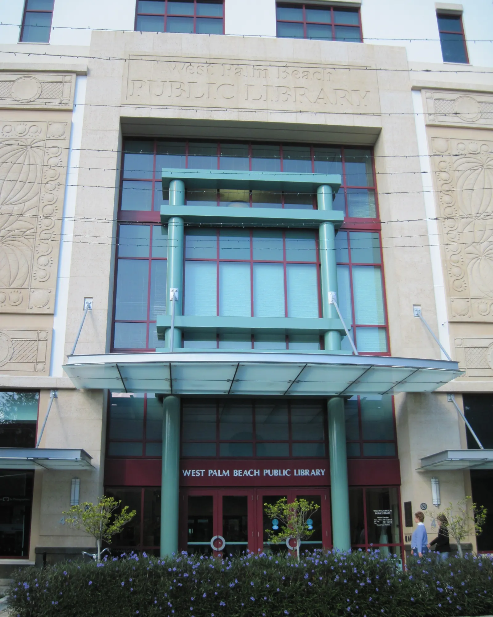 Tall portrait view of the library entrance: a cream stone facade carved with 'West Palm Beach Public Library', a multi-storey glass curtain wall framed in teal steel, a teal entrance canopy and red doors, with a bed of purple flowers in front.
