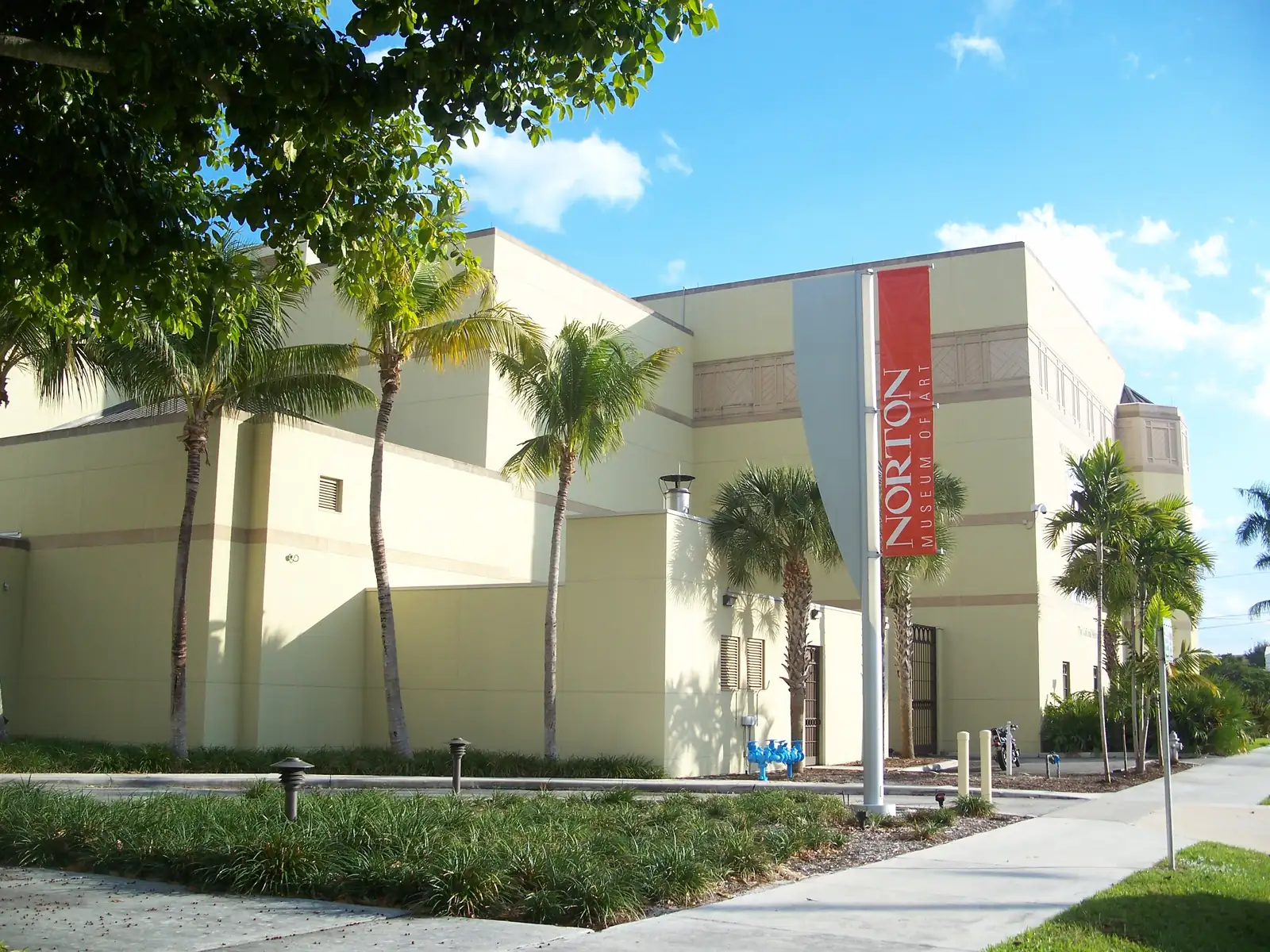 Sunny side view of a cream stucco museum building with stepped walls and small windows, tall palms, a red vertical 'Norton Museum of Art' banner on a pole, and a sidewalk and planted beds under a blue sky with white clouds.