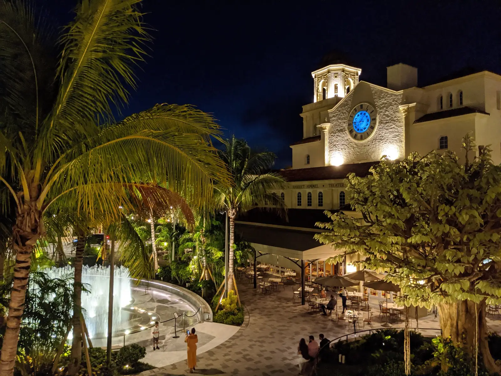 Night view over the CityPlace plaza in 2020: a lit jet fountain among floodlit palms, restaurant tables under umbrellas and the floodlit former church with its clock tower.