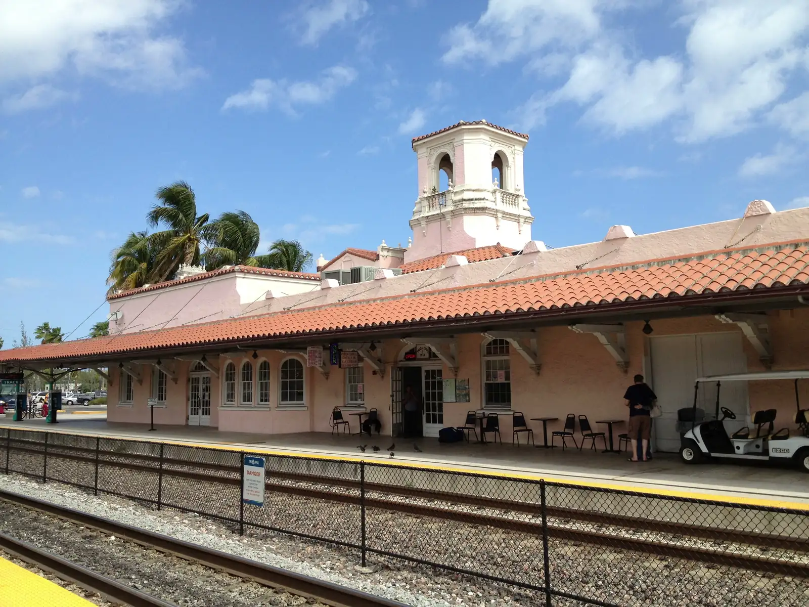 Trackside view of the 1925 Mission-style Seaboard Air Line station: pink stucco walls, a red-tile roof over the platform canopy, a white belfry tower, palms behind, and a fenced track with a yellow-edged platform in the foreground.