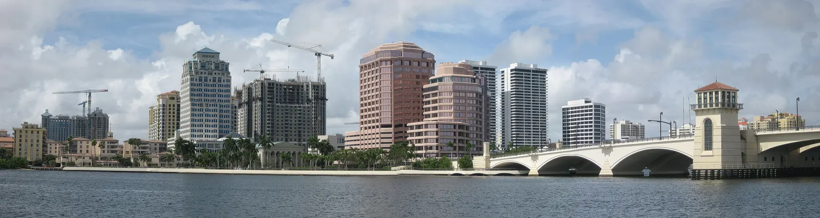 Daytime panorama of the downtown towers across the lagoon under a partly cloudy sky, several still under construction with tower cranes, and at right a white arched drawbridge with a domed bridge-tender tower reaching the shore.