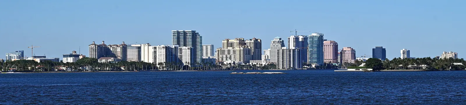 Very wide panorama of the downtown West Palm Beach skyline seen across the deep-blue Lake Worth Lagoon under a clear sky: a long row of mid- and high-rise towers in white, tan, pink and glass-blue, with a low green shoreline of palms and the waterfront below them.