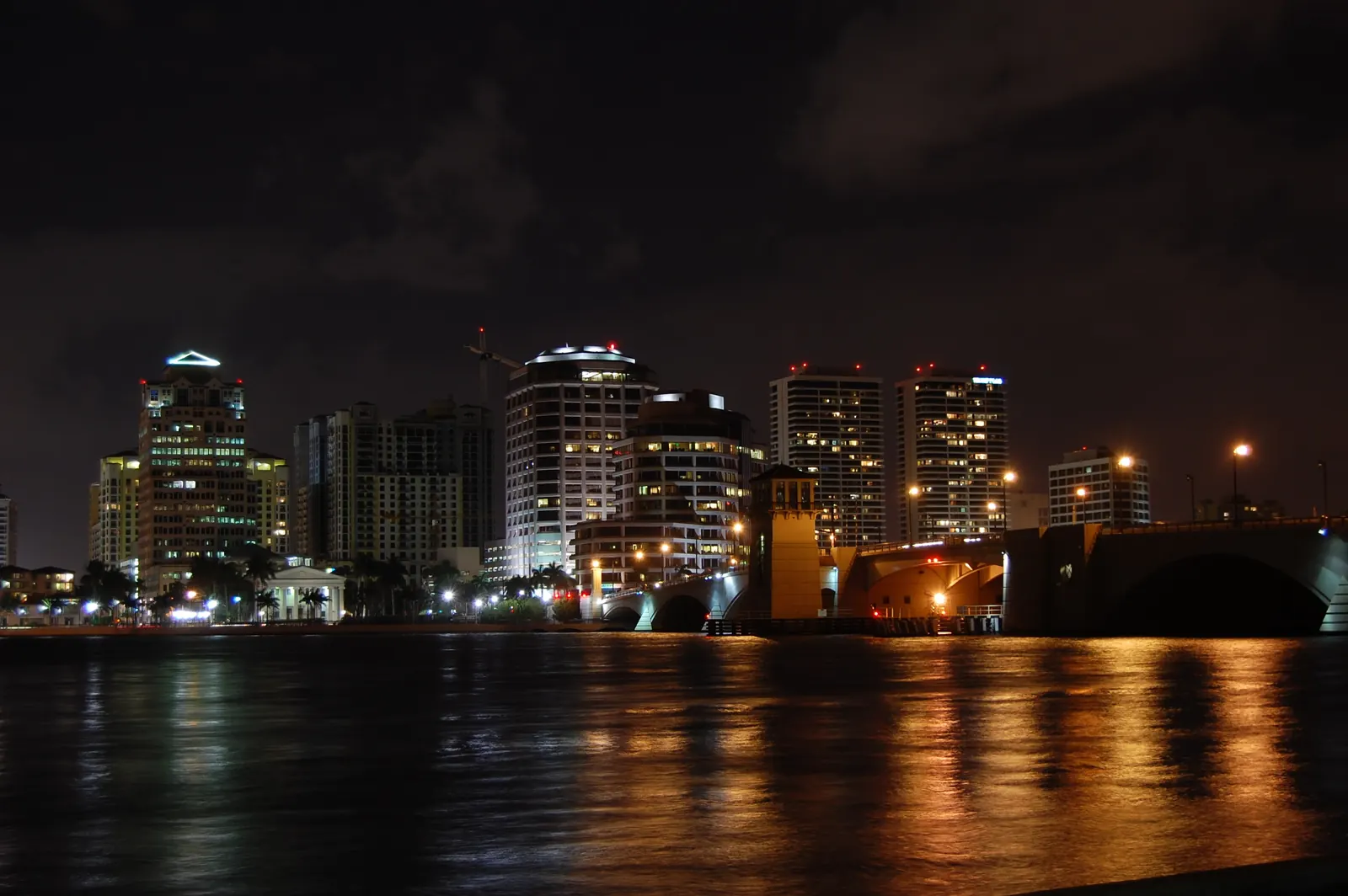 Downtown towers lit up at night across dark water, their lights reflected as orange streaks on the lagoon, with a lit drawbridge and its tender tower at right.