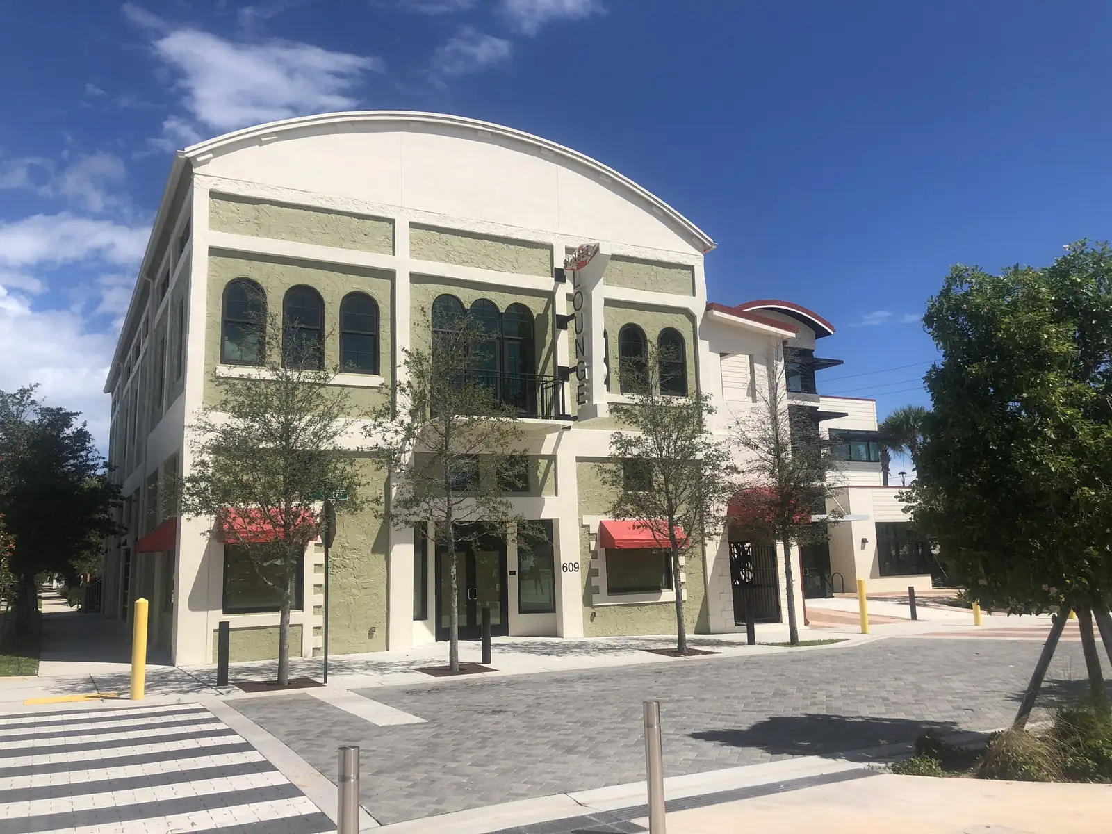 The restored two-storey Sunset Lounge on a corner: sage-green walls with tall arched windows, a curved parapet roof, red awnings, young street trees, bollards and fresh brick paving with a crosswalk under a blue sky with clouds.