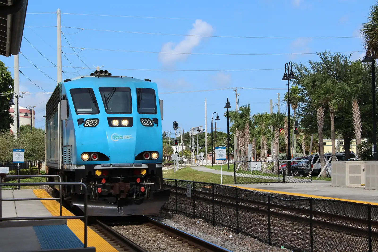 A bright-blue Tri-Rail locomotive with headlights on, approaching along the platform at West Palm Beach station, with signals, lamp posts, palms, parked cars and a fenced track bed under a blue sky with a few clouds.