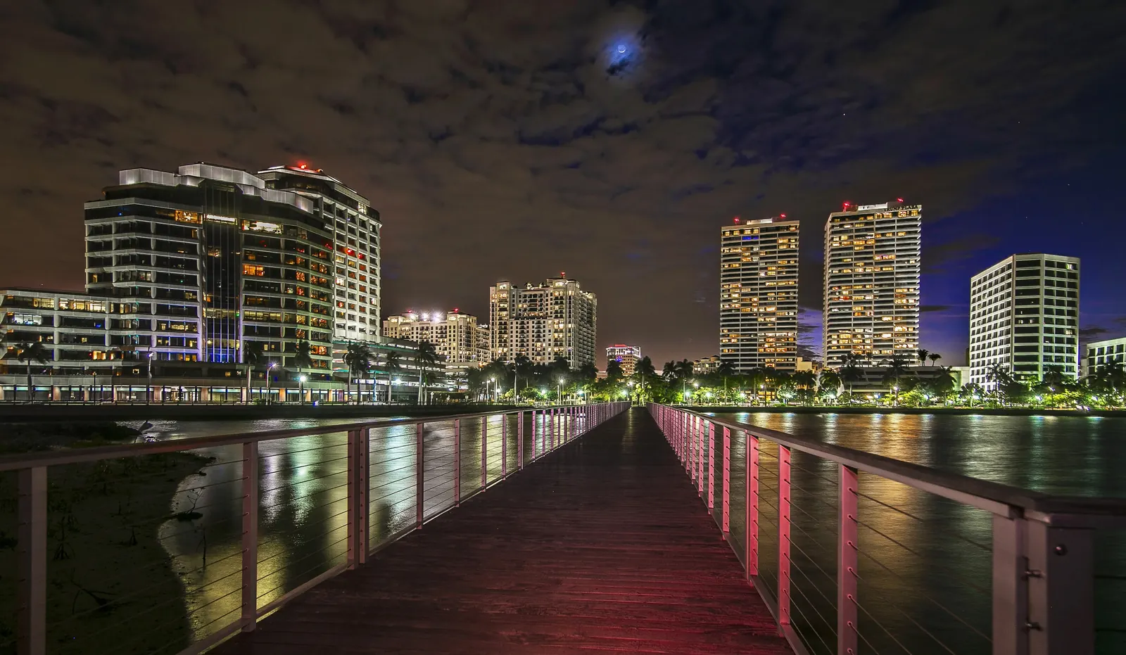 Night view down a long timber pier with lit cable railings reflecting red and white on the deck, leading the eye to the lit downtown waterfront towers, palms and street lights along the shore under a cloudy night sky with a bright moon.