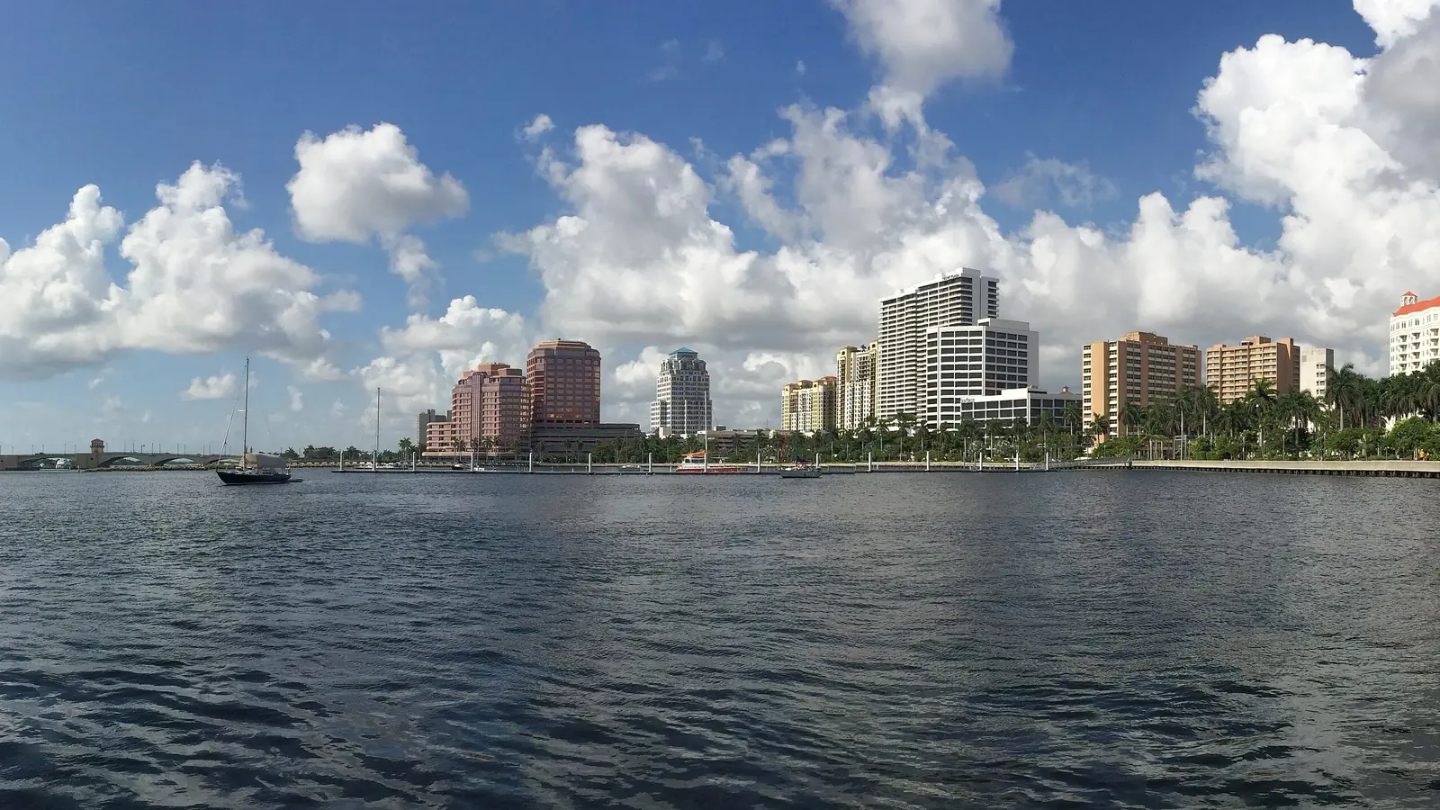 Panorama across choppy blue Intracoastal water to the low downtown skyline and the palm-lined waterfront under a bright sky of big white cumulus clouds, with a single sailboat moored mid-frame.