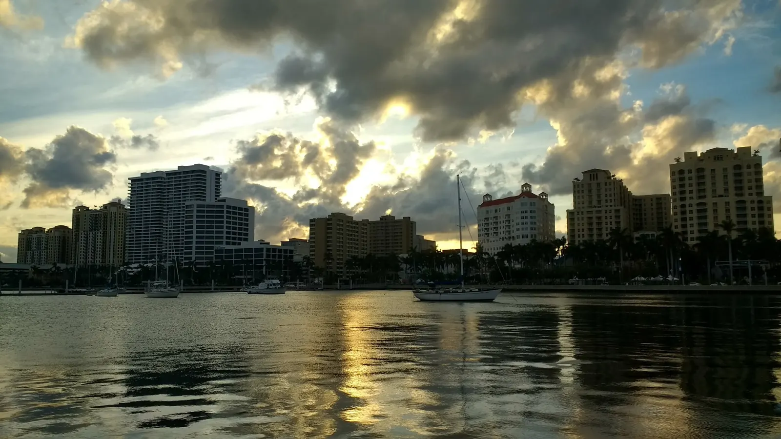 Sunset over the downtown waterfront: dark silhouettes of mid-rise towers and moored sailboats against a dramatic sky of heavy clouds lit gold from behind, mirrored in calm water in the foreground.